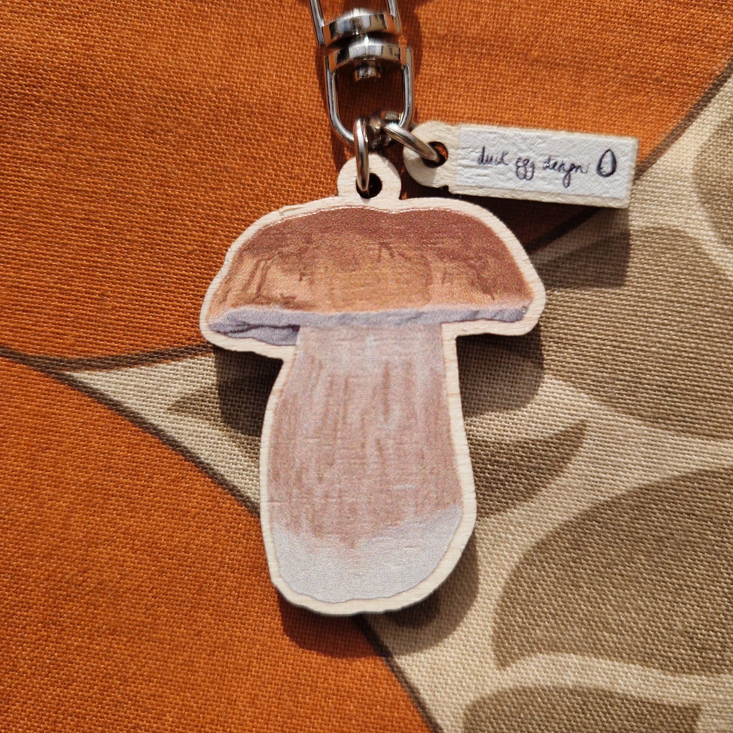 Penny bun orange, brown and white fungi keyring close up showing the fungi and duck egg designs logo charms. The keyring is on a brown and rich orange fabric background.
