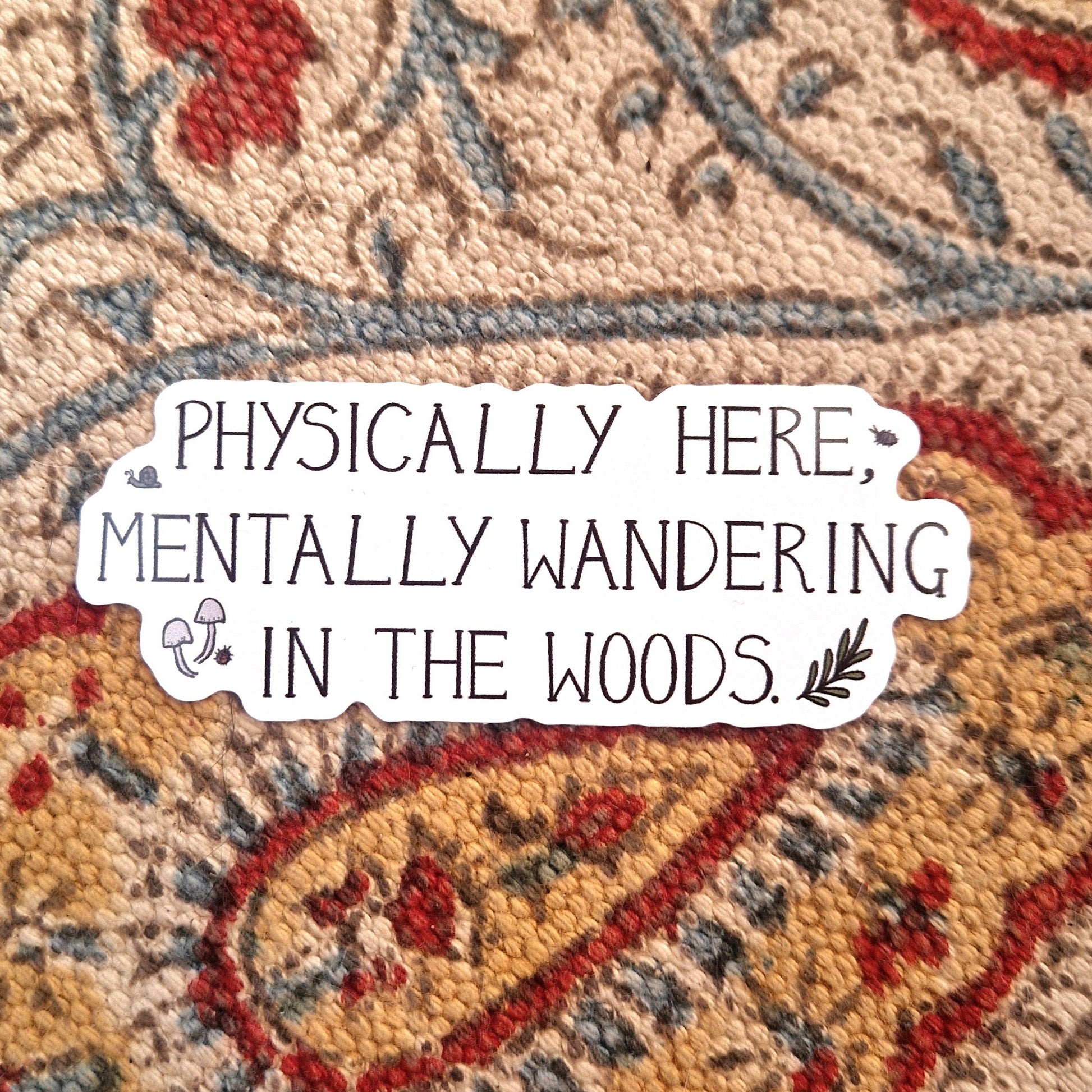 A white sticker featuring the words 'PHYSICALLY HERE, MENTALLY WANDERING IN THE WOODS.' in black handwriting alongside some woodland critters, fungi and a leafy vine. Behind the sticker is a patterned fabric background.