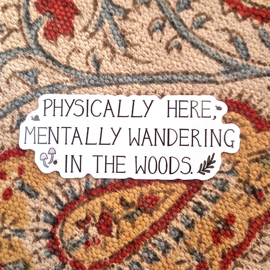 A white sticker featuring the words 'PHYSICALLY HERE, MENTALLY WANDERING IN THE WOODS.' in black handwriting alongside some woodland critters, fungi and a leafy vine. Behind the sticker is a patterned fabric background.