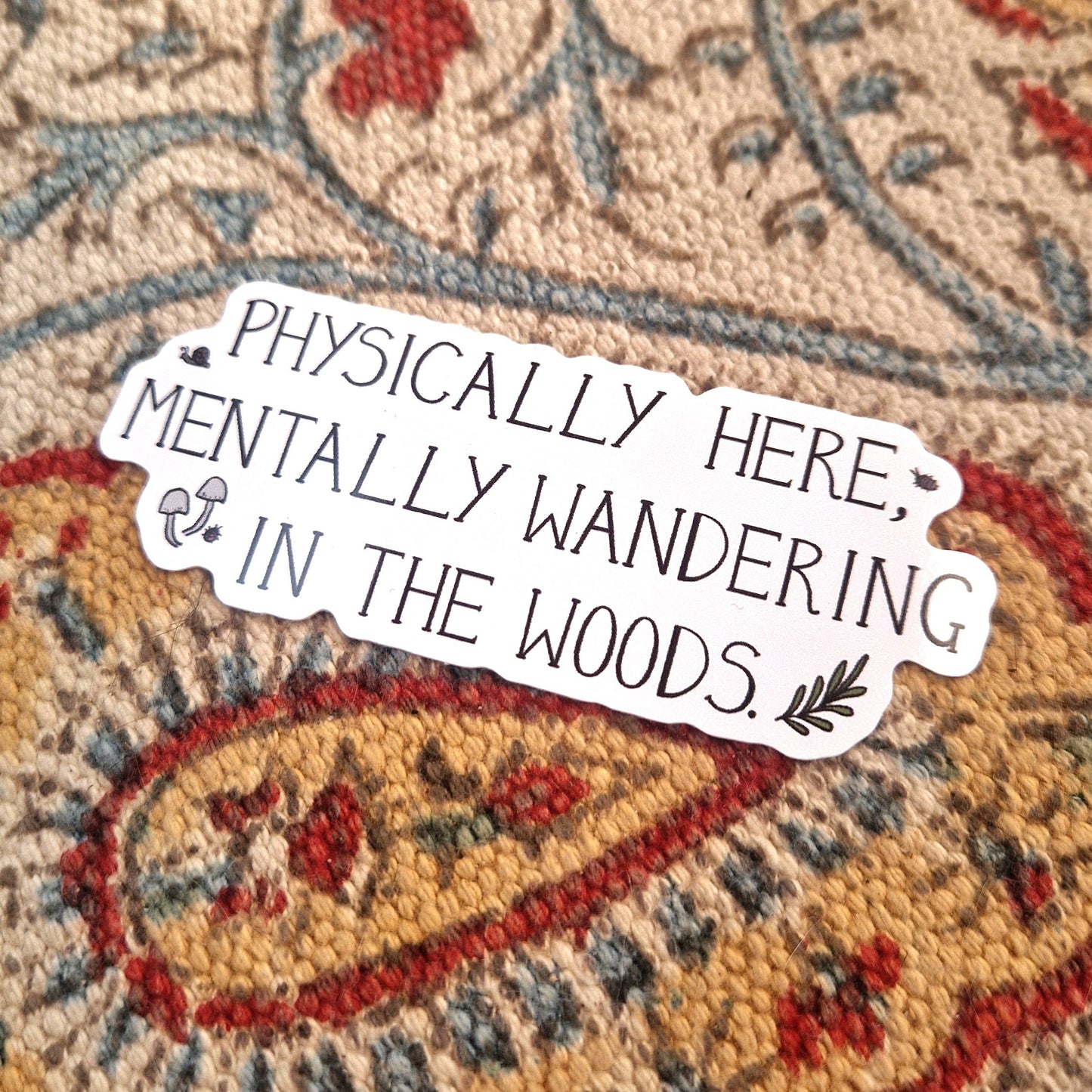 A white sticker featuring the words 'PHYSICALLY HERE, MENTALLY WANDERING IN THE WOODS.' in black handwriting alongside some woodland critters, fungi and a leafy vine. Behind the sticker is a patterned fabric background.