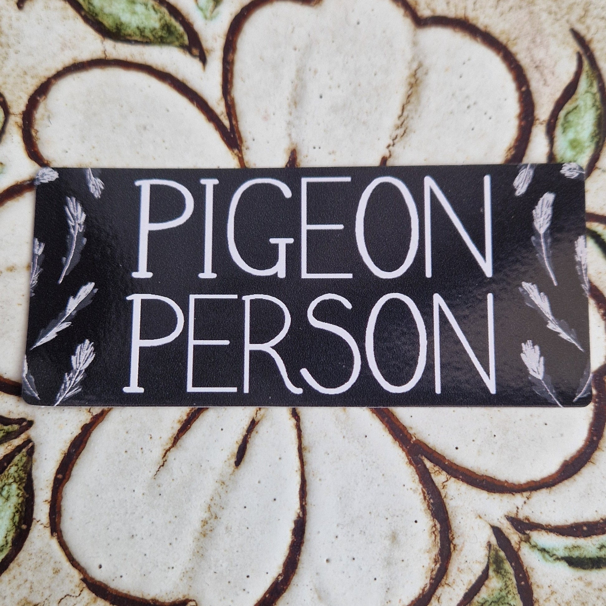 Rectangular sticker with the words 'Pigeon Person' across the middle and grey pigeon feathers around the edge. Behind the sticker is a tile with a flower on it.