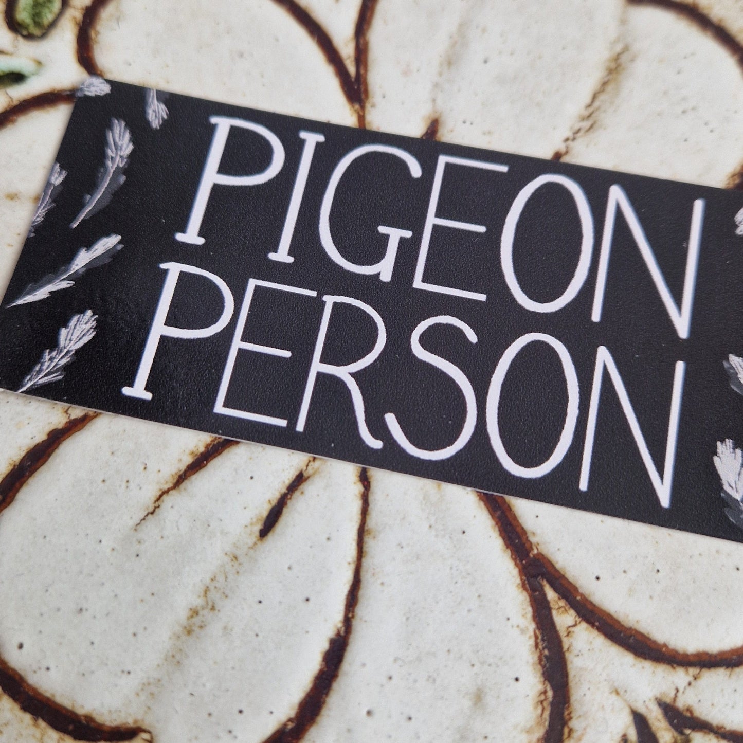A close up of the Pigeon Person sticker showing the pigeon feather design around the edges and the handwritten'Pigeon Person' writing in white. Behind the sticker is a tiled table.