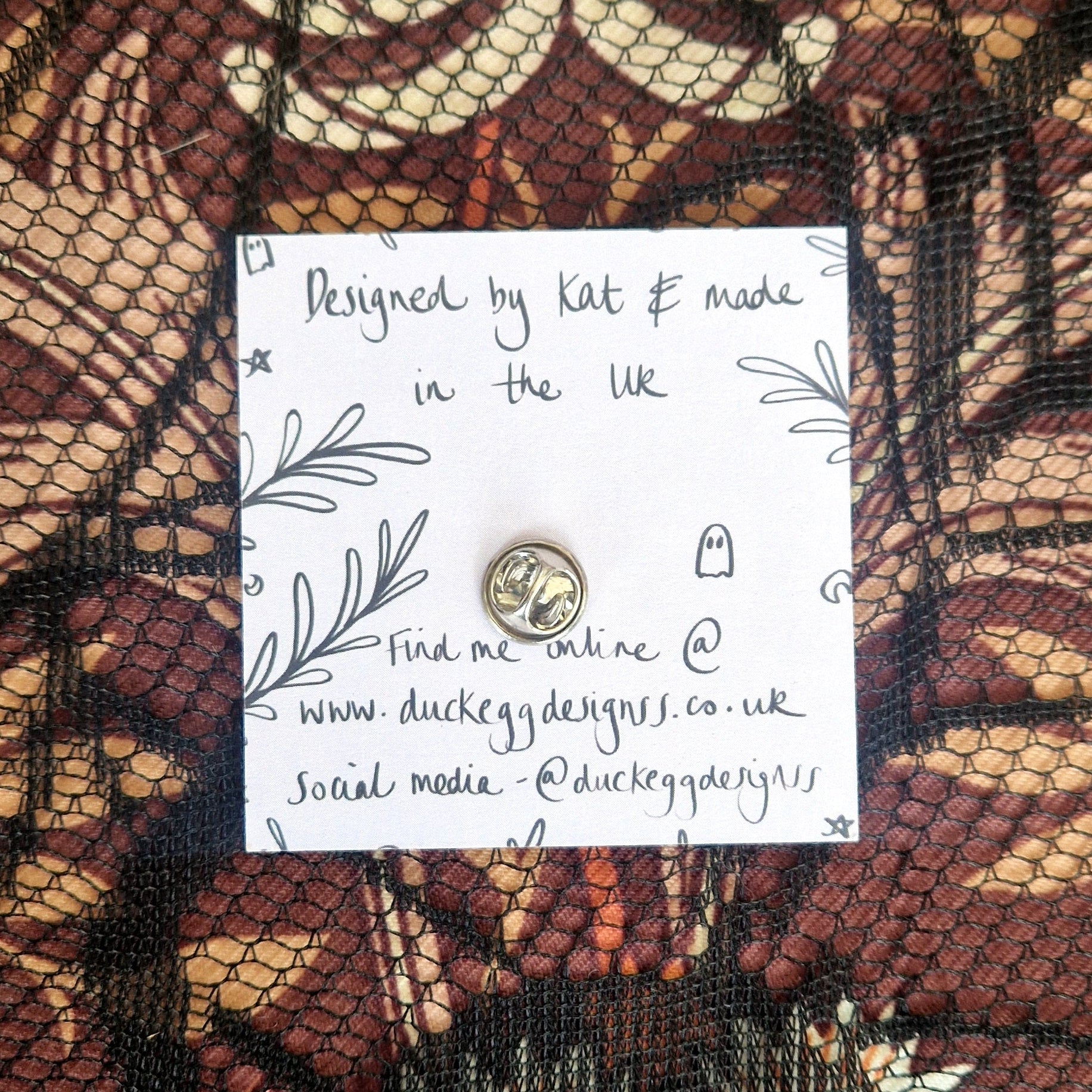 A lilac backing card with a silver butterfly pin back visible sits on a black lace fabric that's on top of a patterned brown fabric.