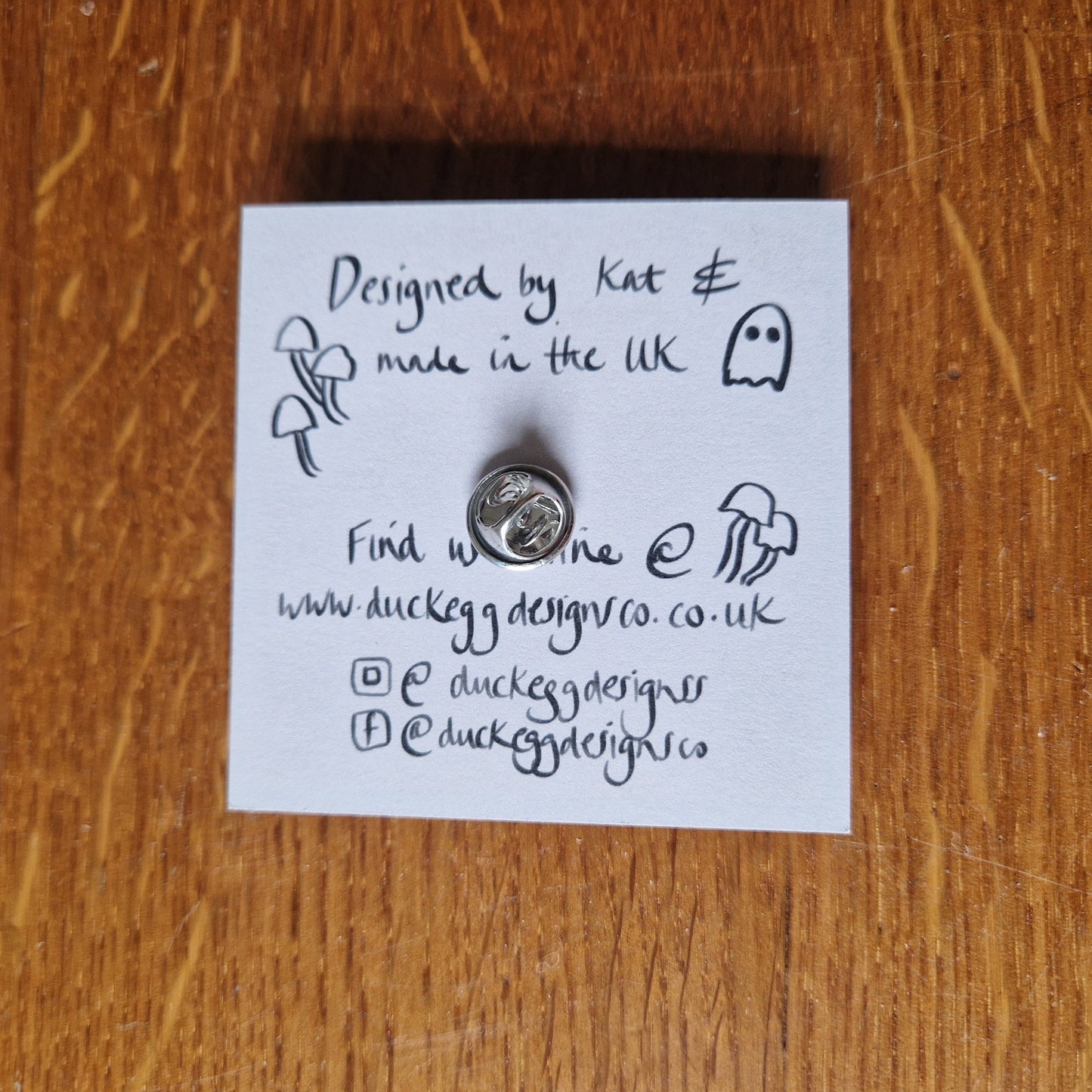 The back of the pin badge showing the silver butterfly clutch back as well as the back of the backing card. Behind the backing card you can see the wooden background.
