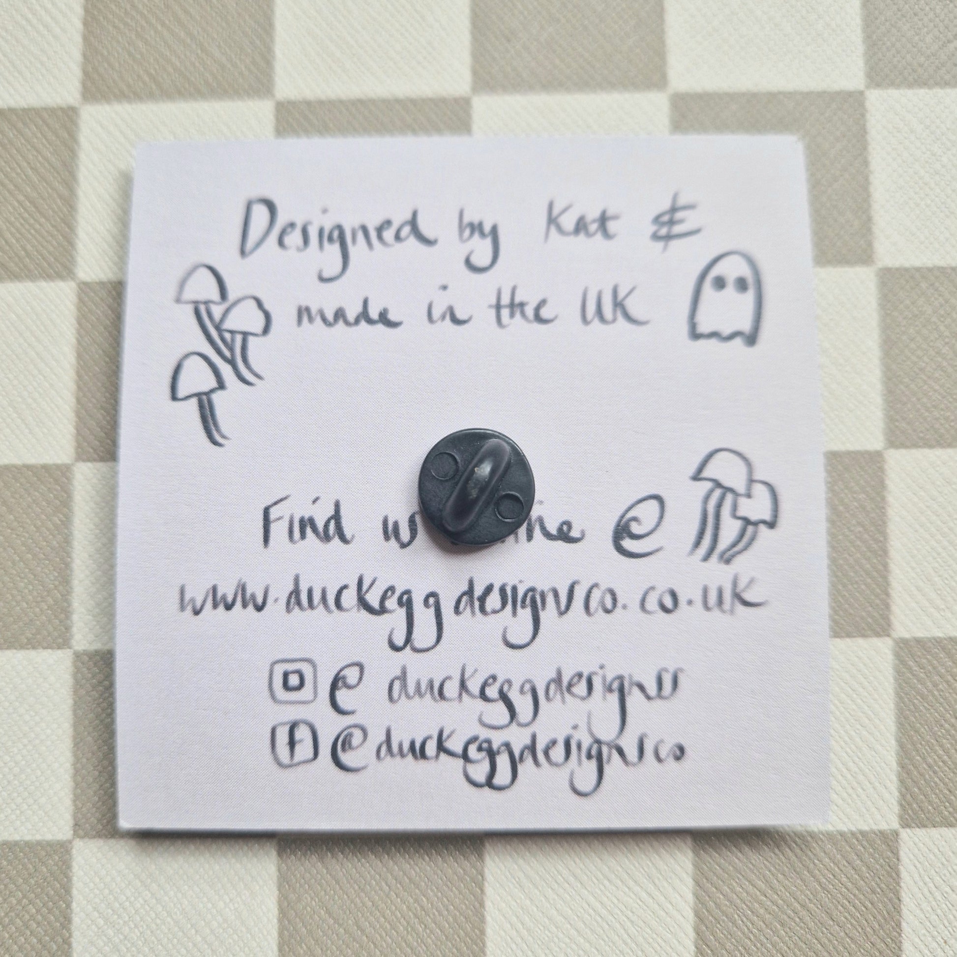 The back of the pin badge showing the rubber back as well as the reverse of the pin badge backing card which is a dusky lilac colour and black handwritten information about the pin.