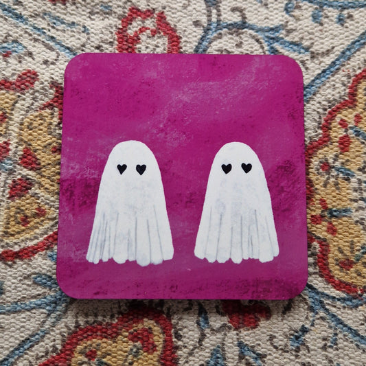 A bright pink square coaster featuring two ghosts with heart eyes sits on a patterned fabric background.
