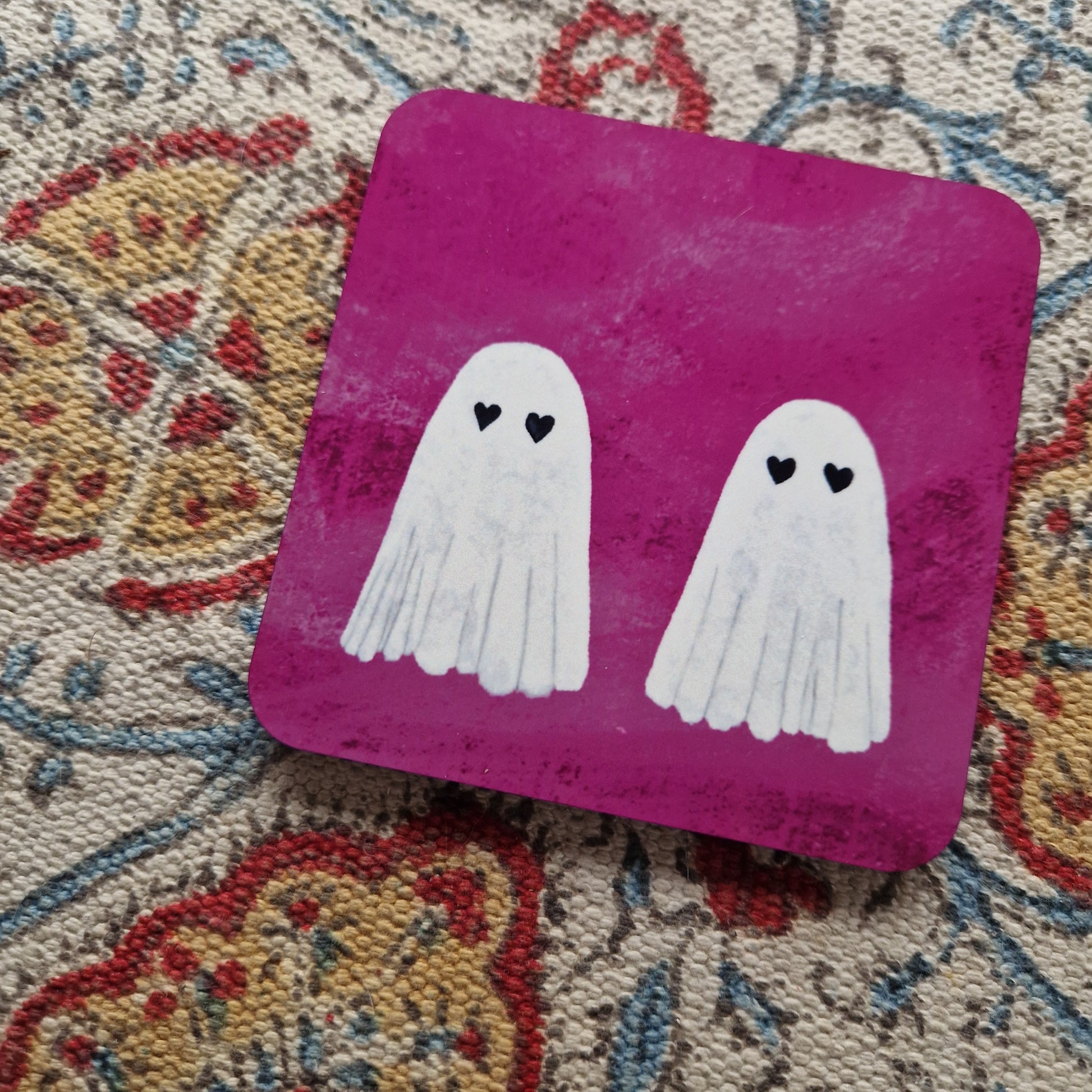 A closer view of the bright pink square coaster, featuring two ghosts with heart eyes. The coaster sits on a patterned fabric background.