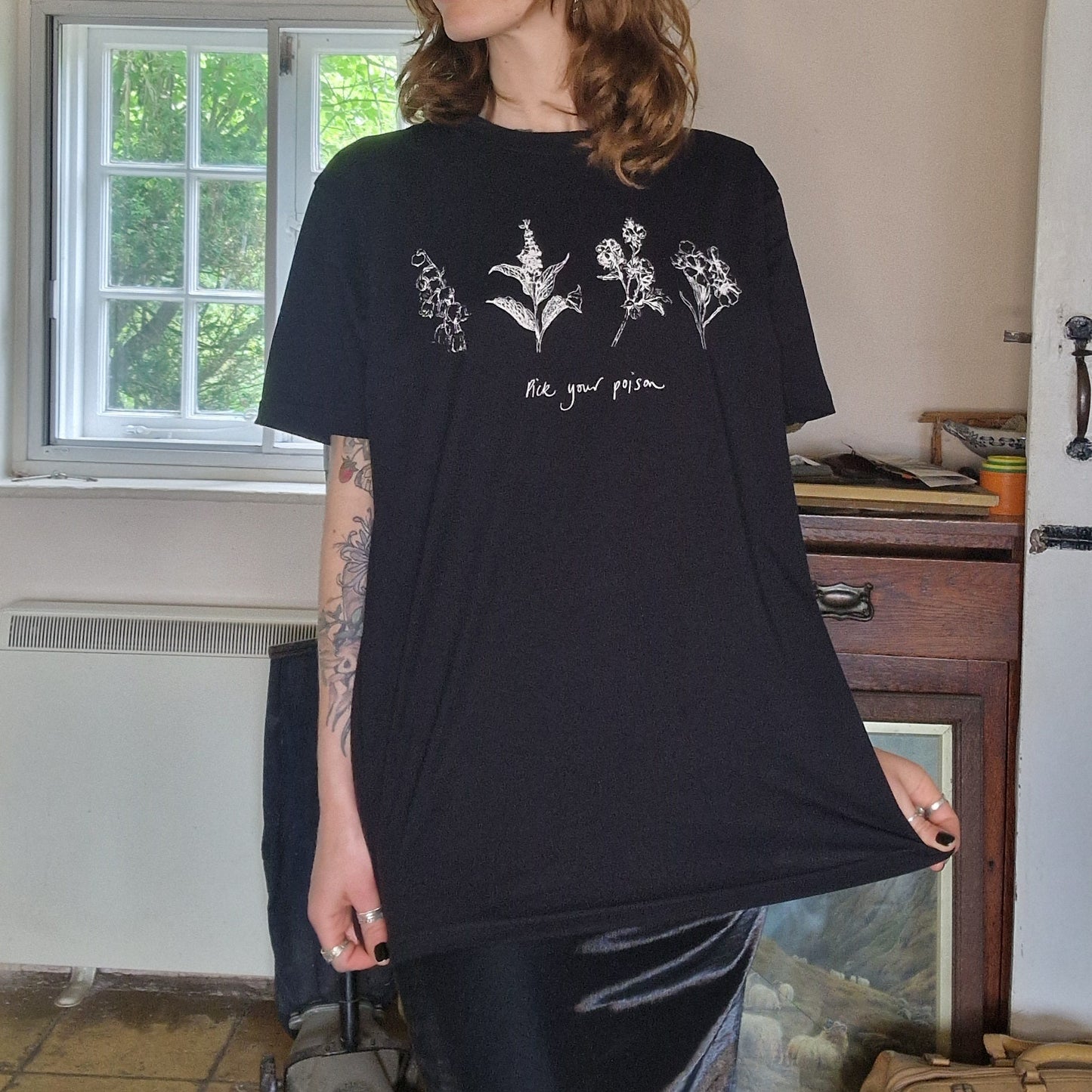 A woman with wavy ginger shoulder length hair wearing a black oversized t-shirt with four poisonous flower across the chest in white with the words 'Pick your poison' underneath. 