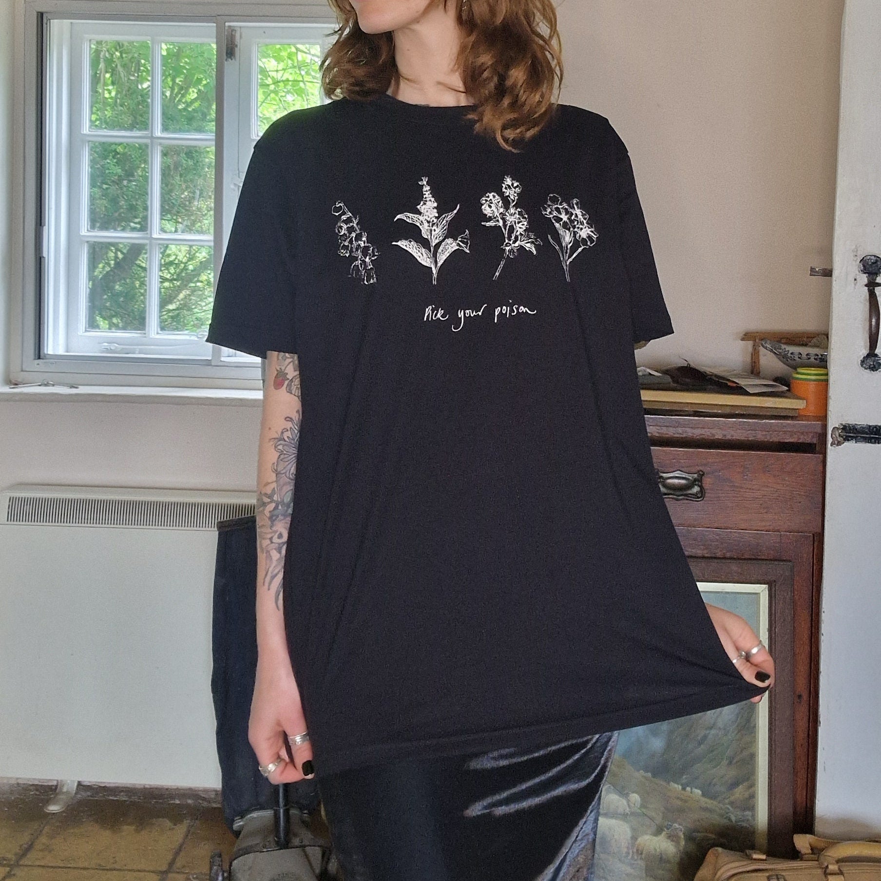 A woman with wavy ginger shoulder length hair wearing a black oversized t-shirt with four poisonous flower across the chest in white with the words 'Pick your poison' underneath. 