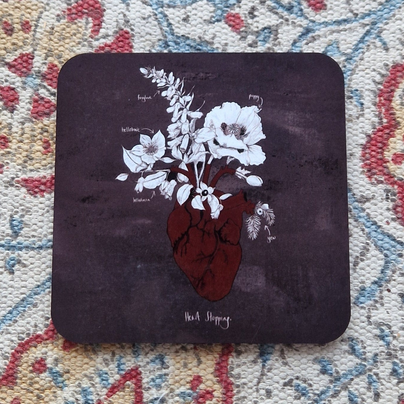 A mottled grey square coaster featuring a red anatomical heart illustration with white and black poison plants growing out from it. Behind the coaster is a patterned fabric background.