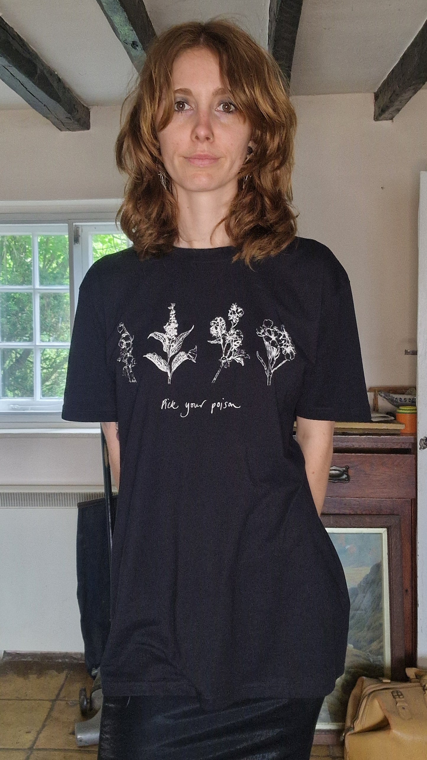 A white woman with wavy auburn hair wearing an oversized black t-shirt with a satin pencil skirt. The t-shirt has white botanical illustrations of plants across the chest with the words 'Pick your poison' written underneath.