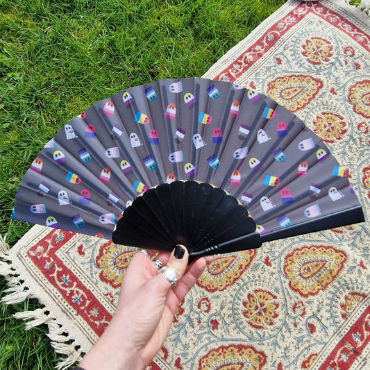A black fan held in front of grass and a patterned rug. The fan has a repeating print of the different pride flags as ghosts.