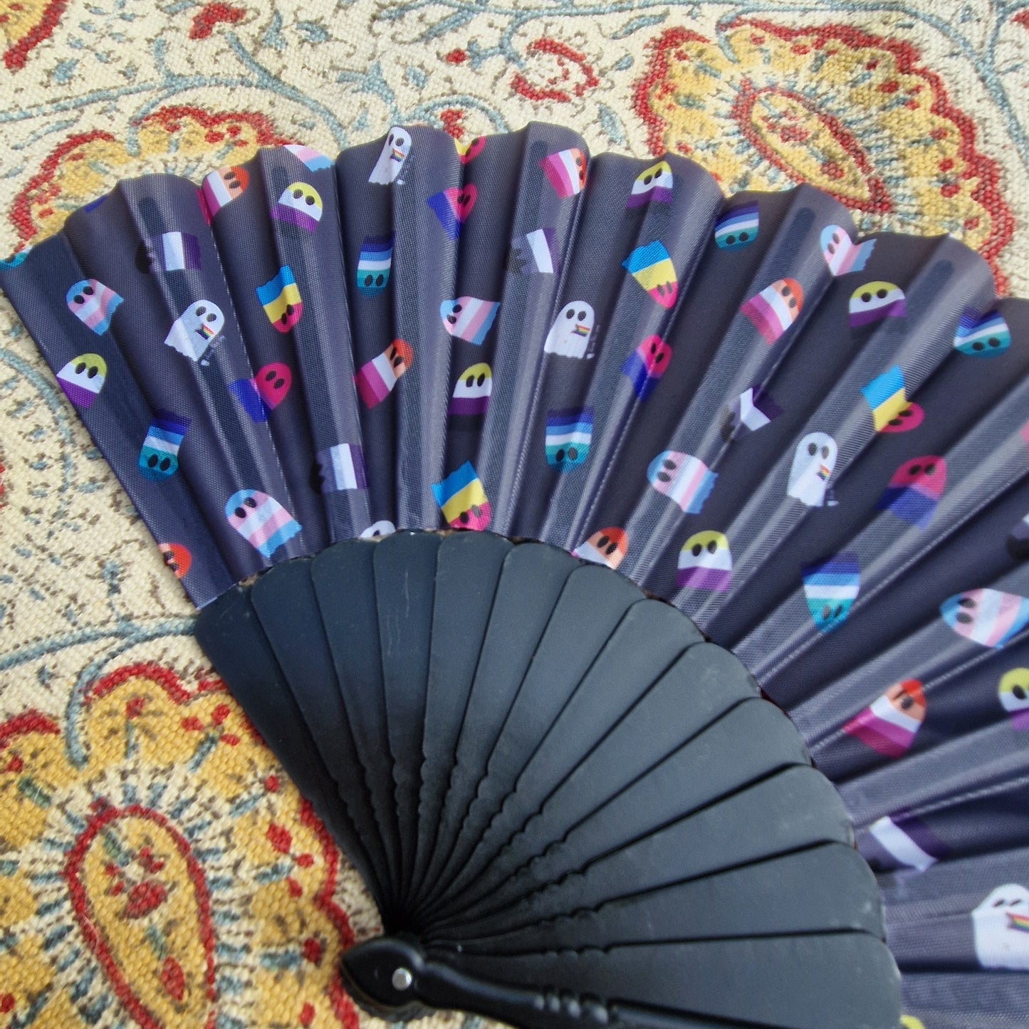 A close up of the black fan showing the detail in the pride flag ghosts repeating print. The fan rests on a patterned rug. 