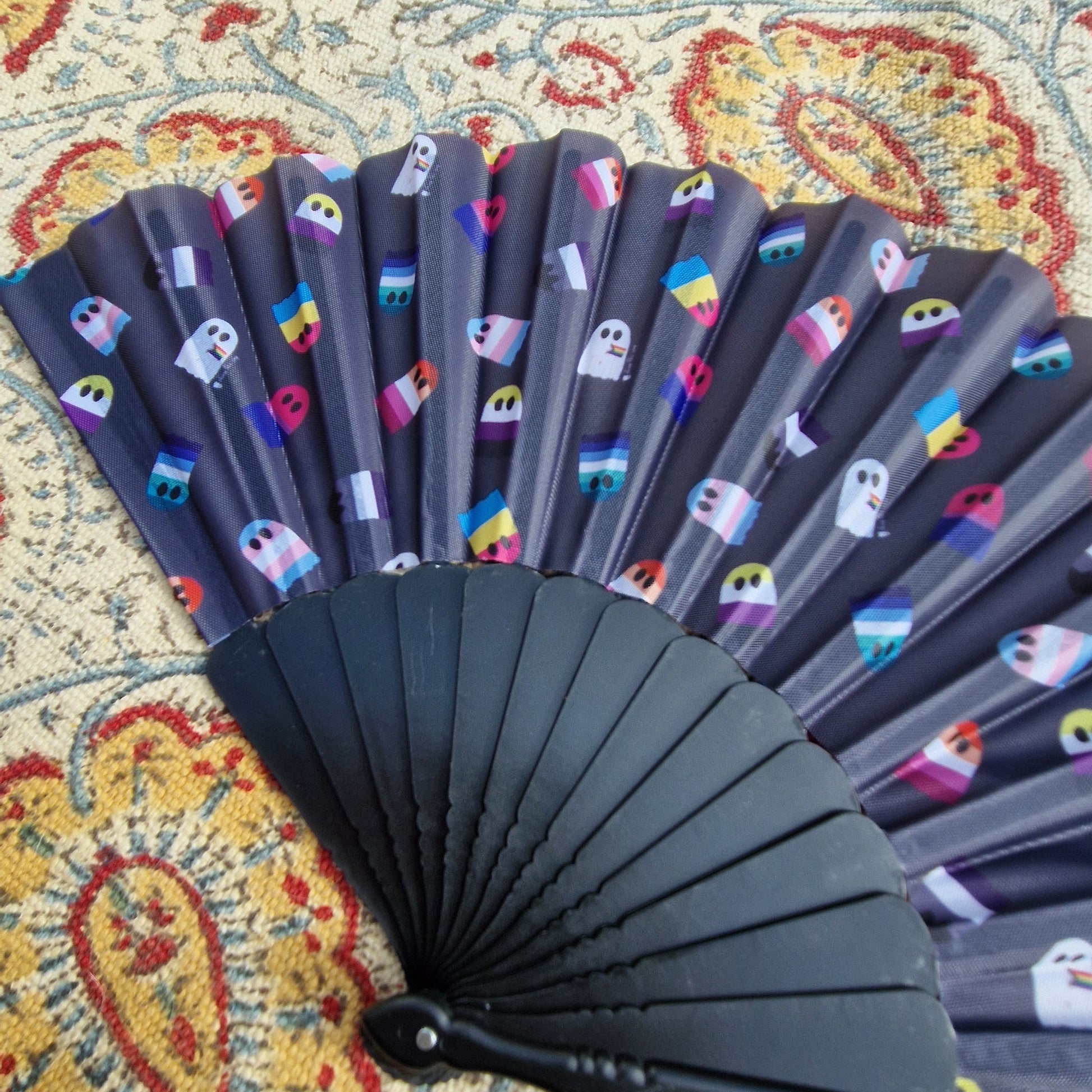 A close up of the black fan showing the detail in the pride flag ghosts repeating print. The fan rests on a patterned rug. 