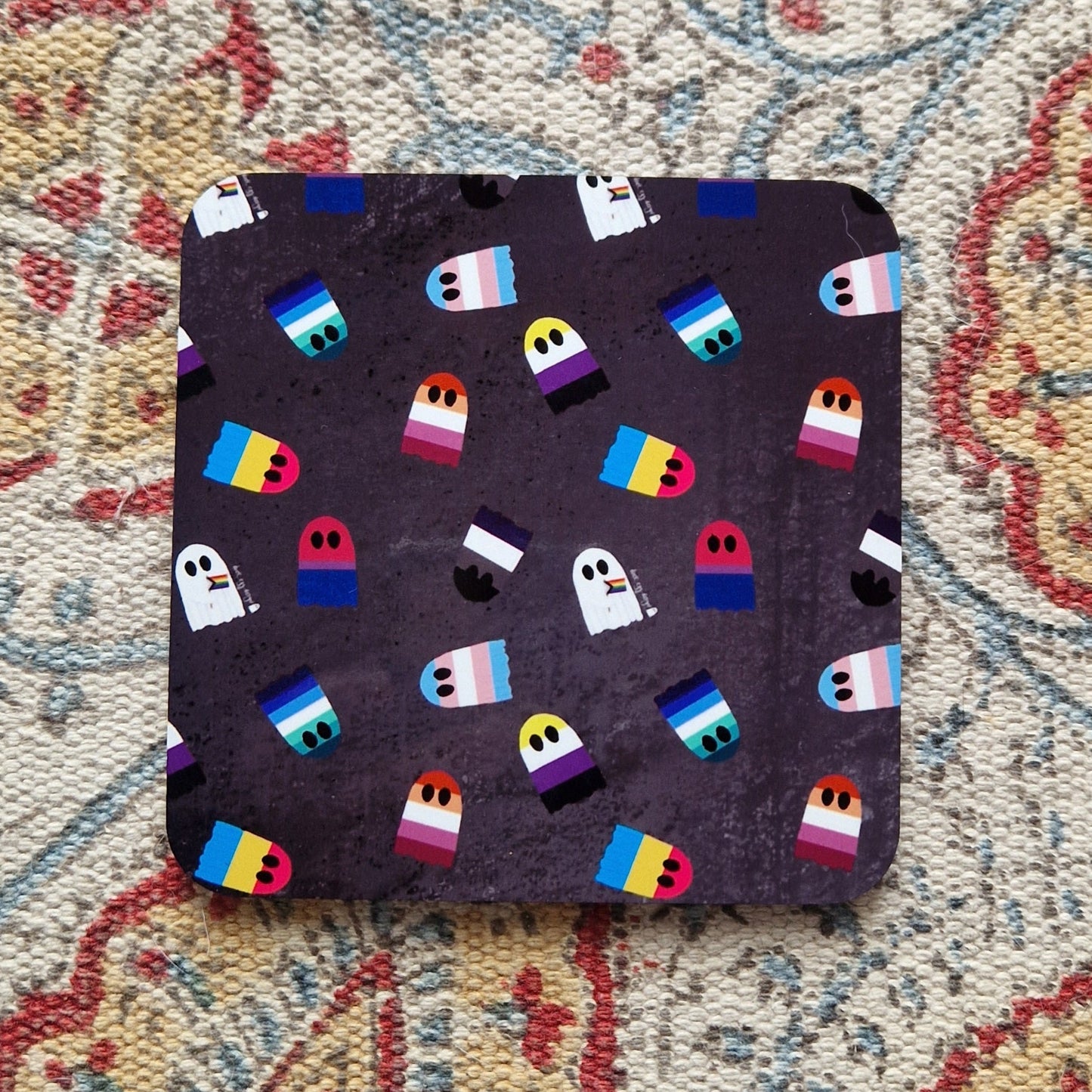A grey square coaster with different pride flags as ghosts sits on a patterned fabric background.