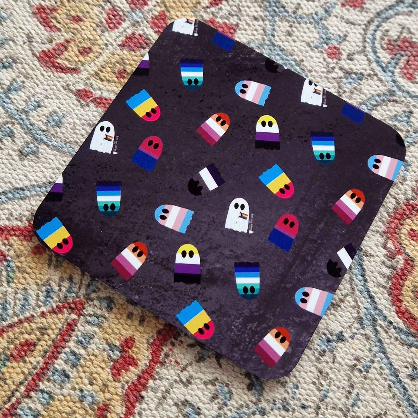 A grey square coaster with different pride flags as ghosts sits on a patterned fabric background.