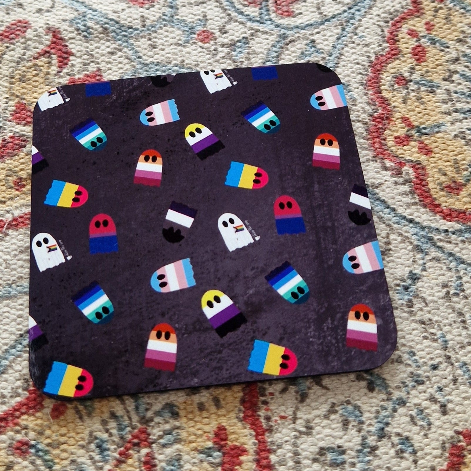 A grey square coaster with different pride flags as ghosts sits on a patterned fabric background.