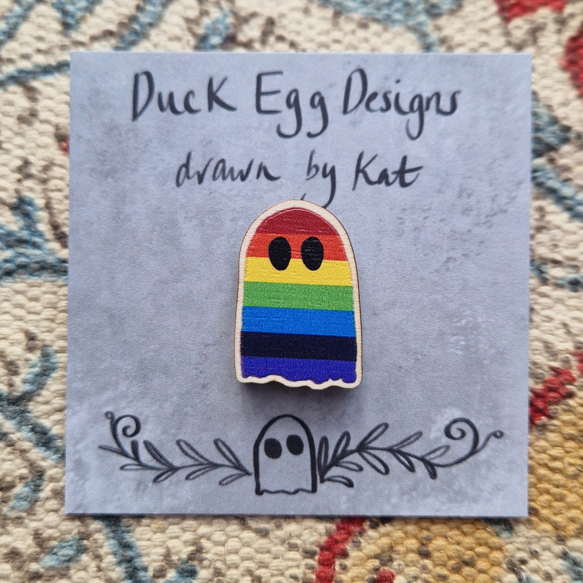 A wooden pin badge in the shape of a ghost in rainbow colours sits on a grey backing card that has the words 'Duck Egg Designs drawn by Kat' in black writing.