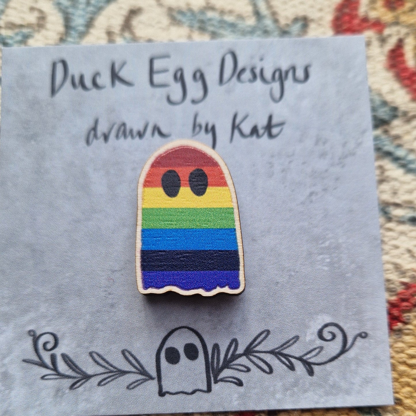 A closer view of the rainbow ghost pin badge showing the wooden texture of the pin. The pid badge sits on a grey backing card with the words 'Duck Egg Designs drawn by Kat' across the top and a ghost and leafy tendrils design across the bottom.