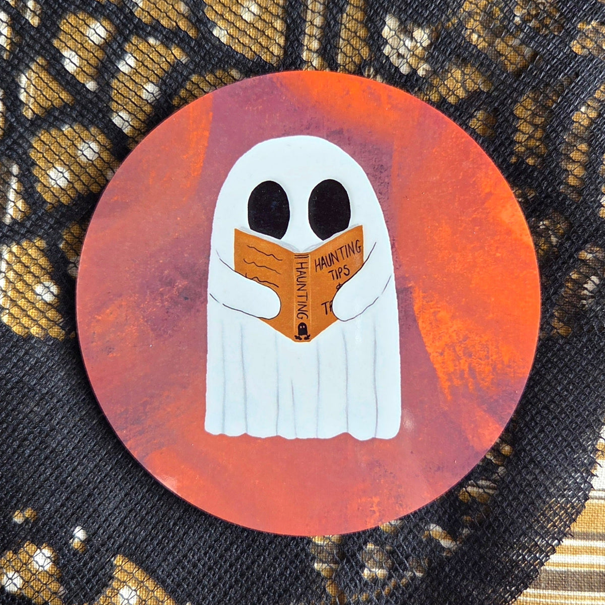 A warm orangey pink round coaster with a white ghost reading an orange book with the title haunting tips. Behind the coaster is a brown patterned fabric with a black lace overlay. 