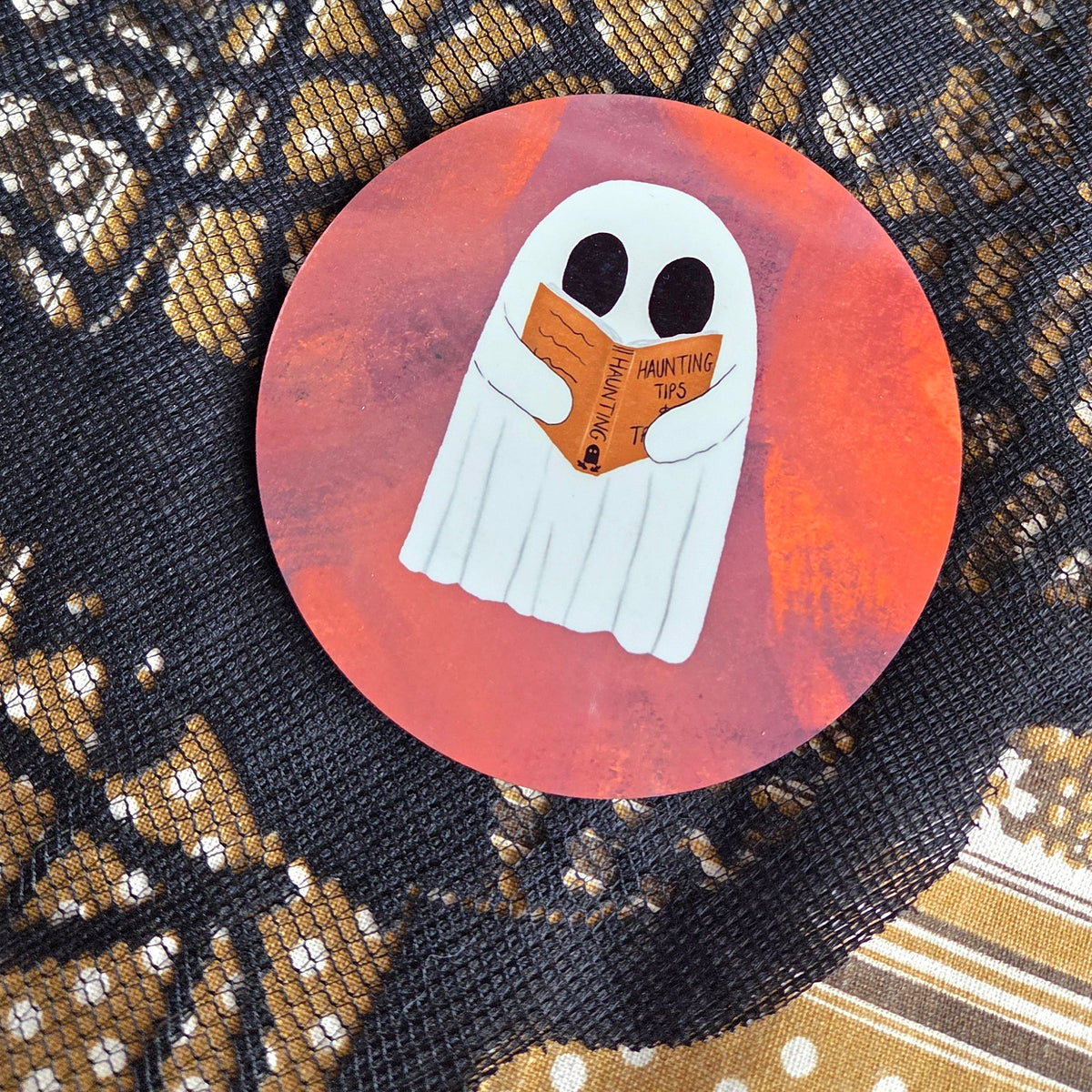 A warm orangey pink round coaster with a white ghost reading an orange book with the title haunting tips. Behind the coaster is a brown patterned fabric with a black lace overlay. 