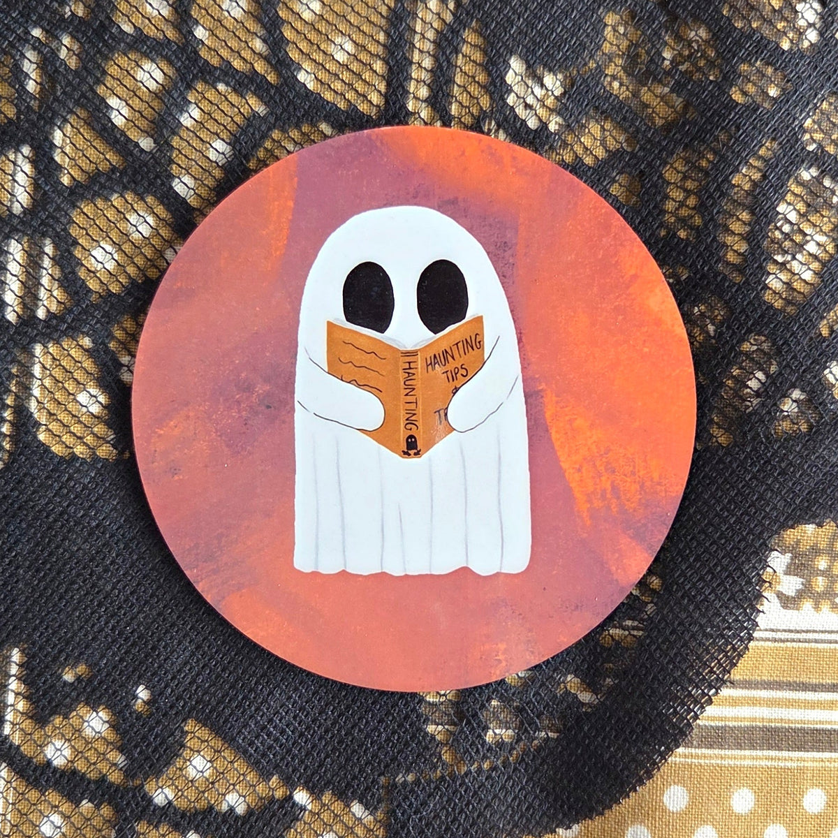 A warm orangey pink round coaster with a white ghost reading an orange book with the title haunting tips. Behind the coaster is a brown patterned fabric with a black lace overlay. 