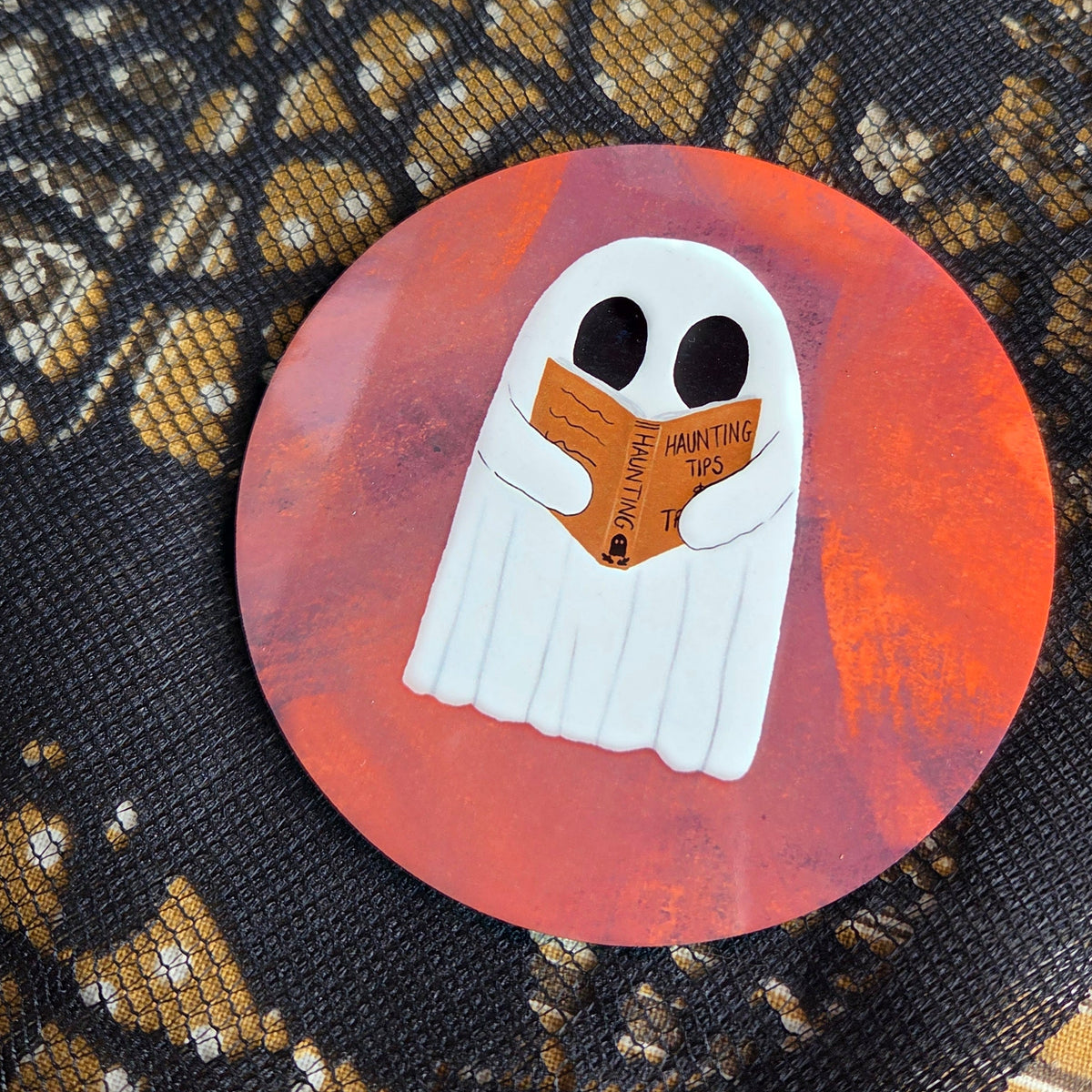 A warm orangey pink round coaster with a white ghost reading an orange book with the title haunting tips. Behind the coaster is a brown patterned fabric with a black lace overlay. 
