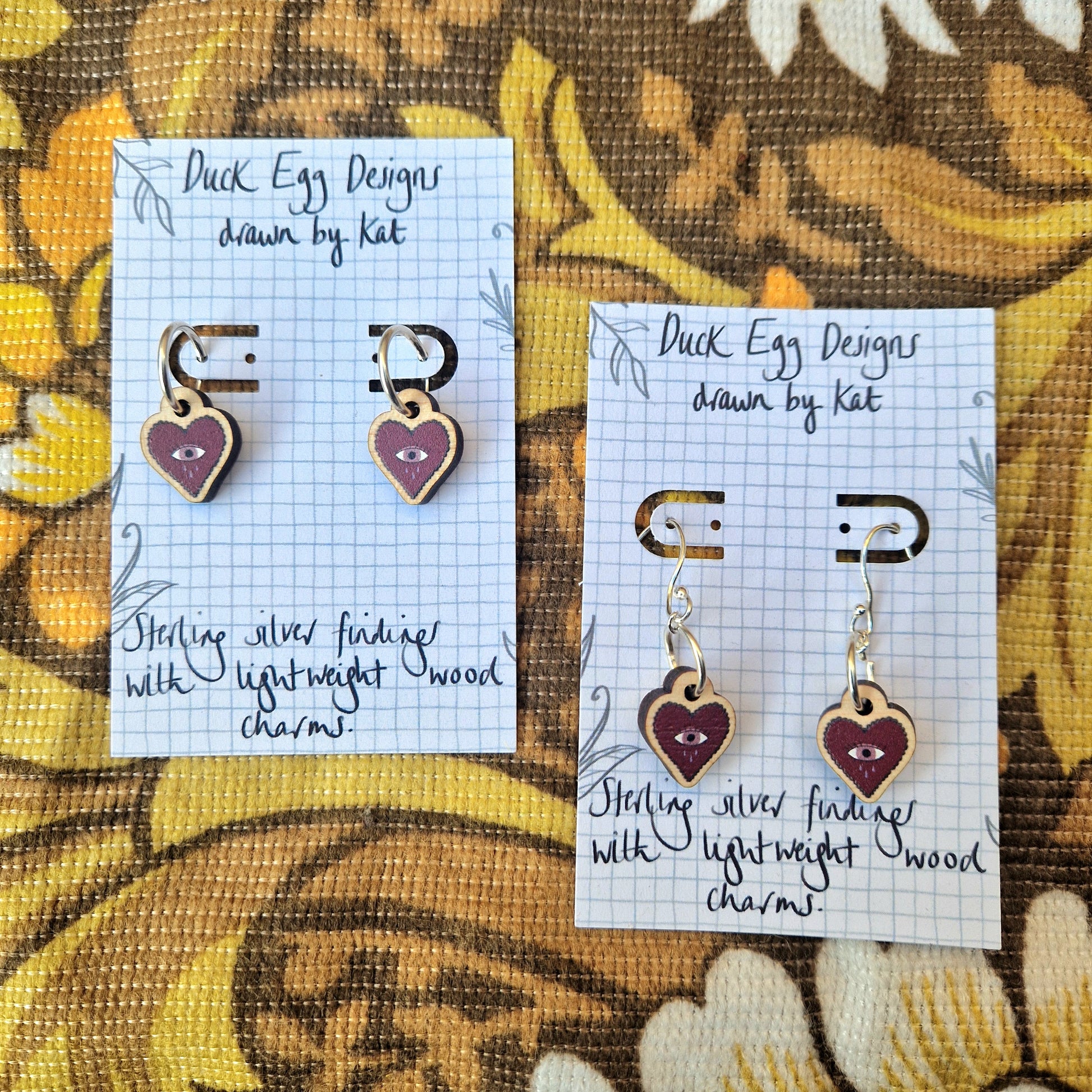 Two pairs of red heart earrings on white backing cards. The hearts have an dusky pink eye in the middle with three blue teardrops falling from it. The heart charms are on hoops on the left and shepherds crooks on the right. Behind the backing cards are a vintage floral brown and orange fabric.
