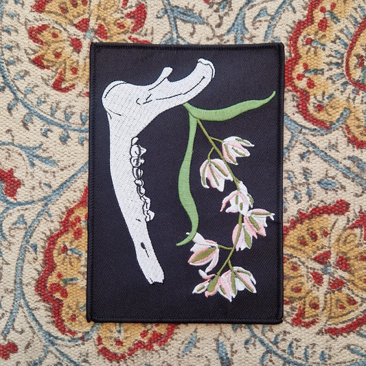 A black rectangular patch sits on a patterned fabric background.  The patch featured a deer jawbone with a green, pink and white orchid plant growing from it.