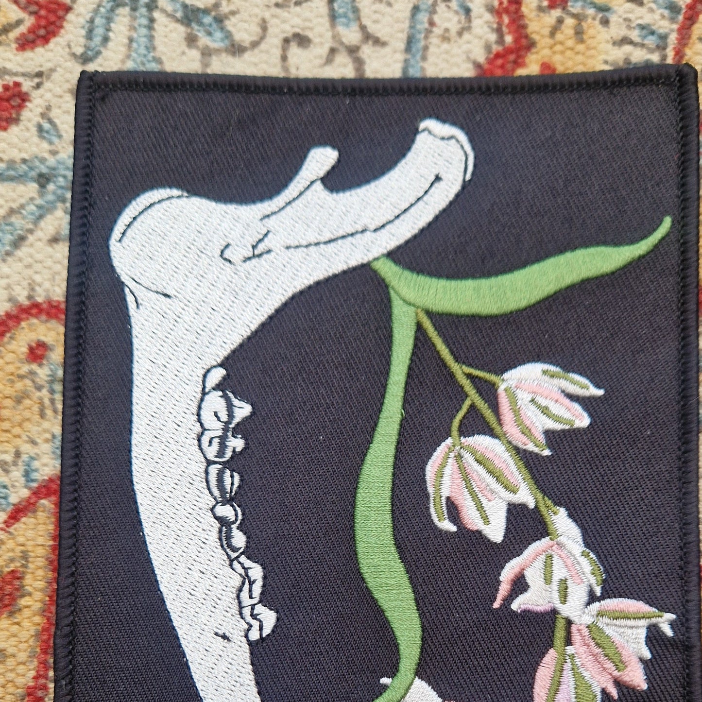 A  close up of the black rectangular patch sits on a patterned fabric background.  The patch features a deer jawbone with a green, pink and white orchid plant growing from it.