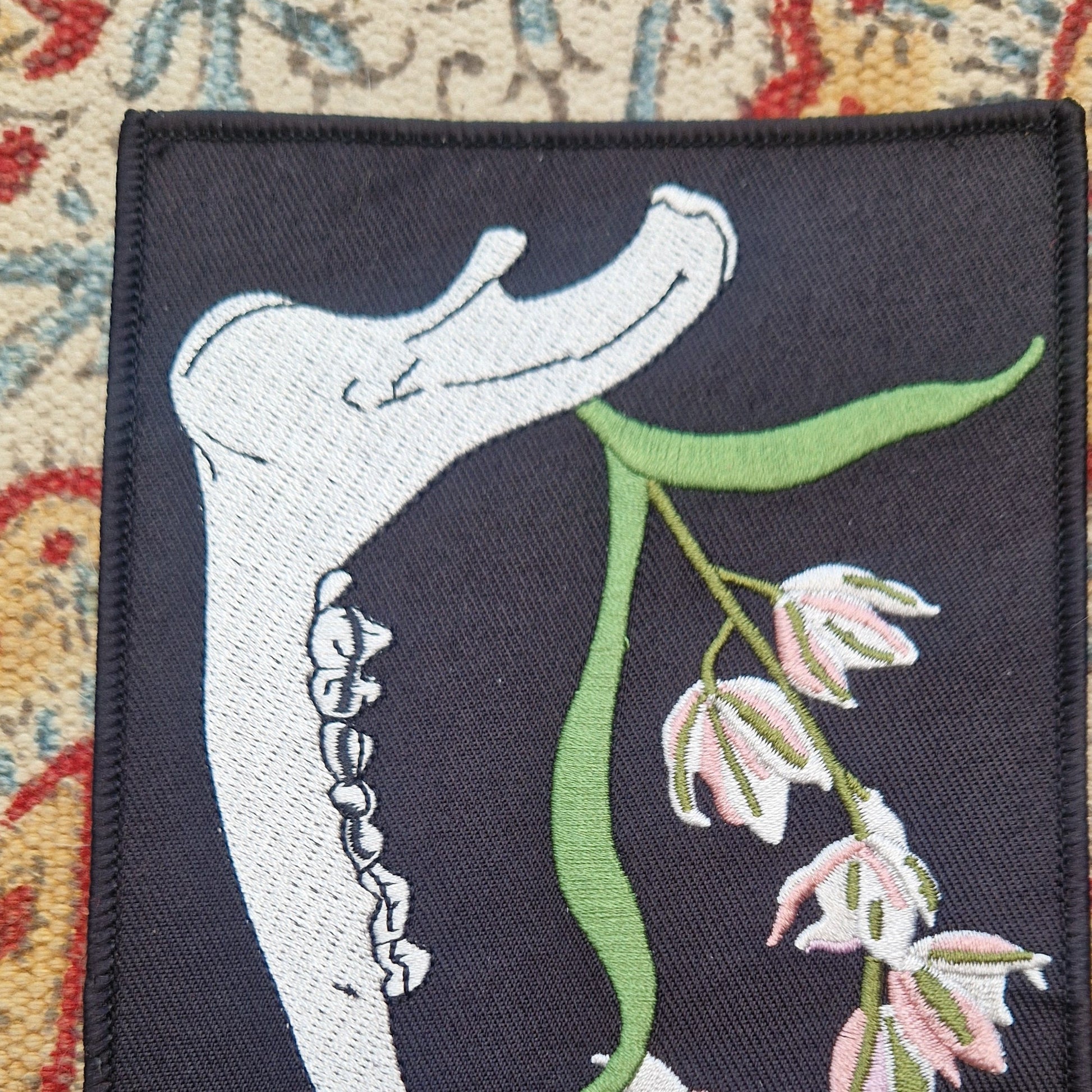 A  close up of the black rectangular patch sits on a patterned fabric background.  The patch features a deer jawbone with a green, pink and white orchid plant growing from it.