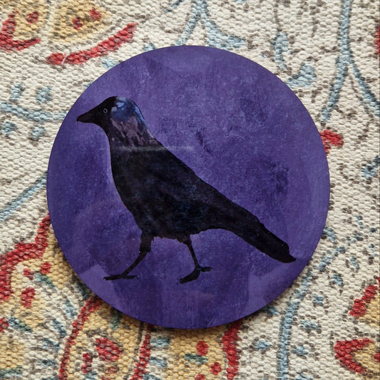 A round purple coaster with a black jackdaw design sits on a patterned white fabric background.