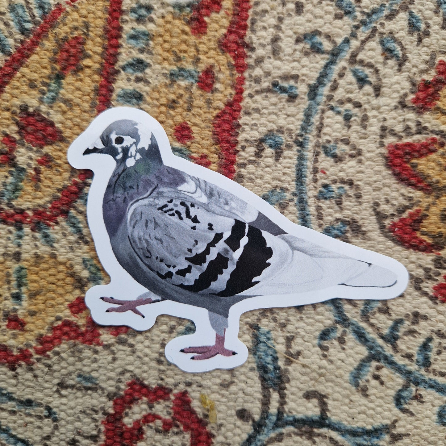 A sticker of a grey, white and black pigeon with pink feet sits on a patterned fabric background.