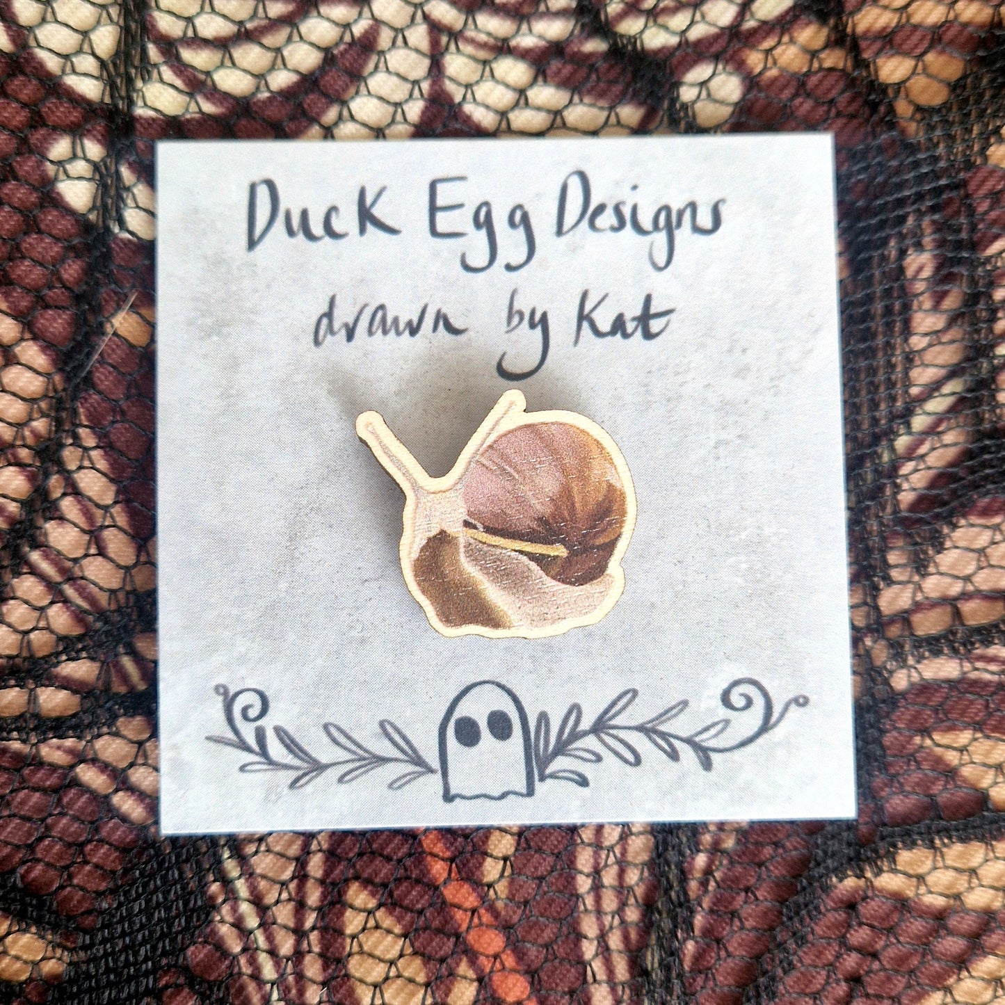 A snail pin badge printed onto eco-friendly wood, on a grey backing card with the words 'Duck Egg Designs drawn by Kat' across the top. Behind the backing card is a black lace fabric on top of a brown patterned fabric.