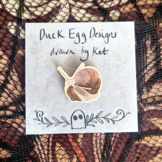 A snail pin badge printed onto eco-friendly wood, on a grey backing card with the words 'Duck Egg Designs drawn by Kat' across the top. Behind the backing card is a black lace fabric on top of a brown patterned fabric.