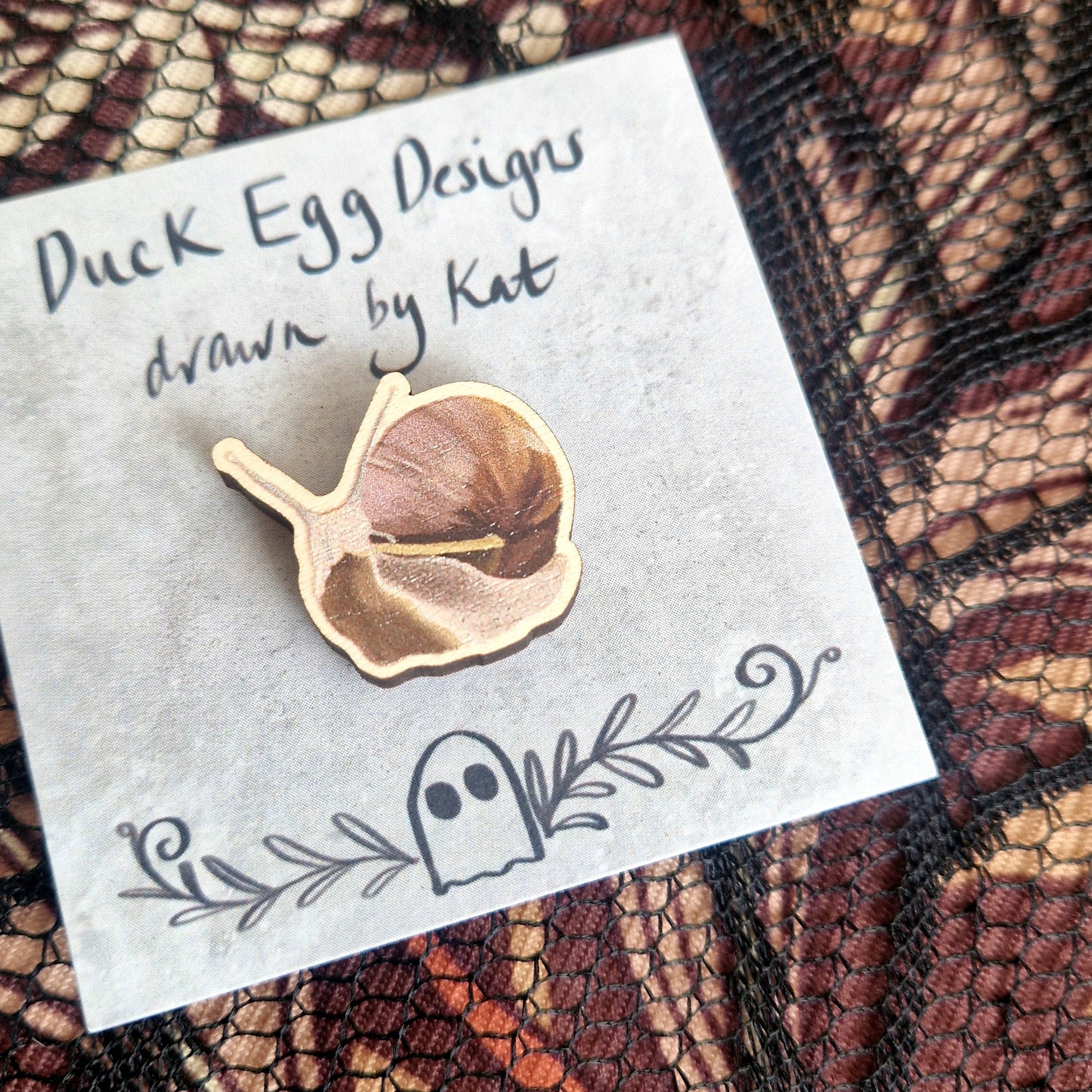 A snail pin badge printed onto eco-friendly wood, on a grey backing card with the words 'Duck Egg Designs drawn by Kat' across the top. Behind the backing card is a black lace fabric on top of a brown patterned fabric.