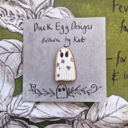 A white ghost pin with a black spiderweb design sits on a grey backing card with the words ' Duck Egg Designs drawn by Kat' in black handwriting.  Behind the backing card is a rich green fabric with a white and black plant design.