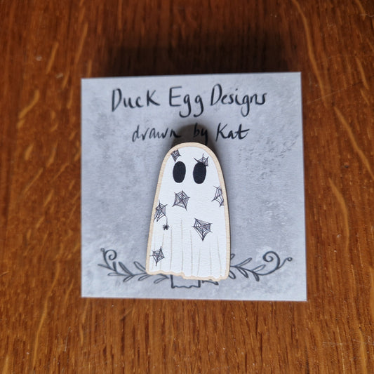 A spiderweb covered ghost pin badge sits on a grey backing card with the words 'Duck Egg Designs drawn by Kat' across the top. Behind the card you can see a wooden background.