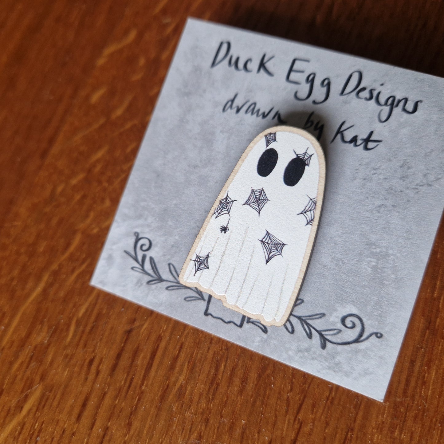 A spiderweb covered ghost pin badge sits on a grey backing card with the words 'Duck Egg Designs drawn by Kat' across the top. Behind the card you can see a wooden background.
