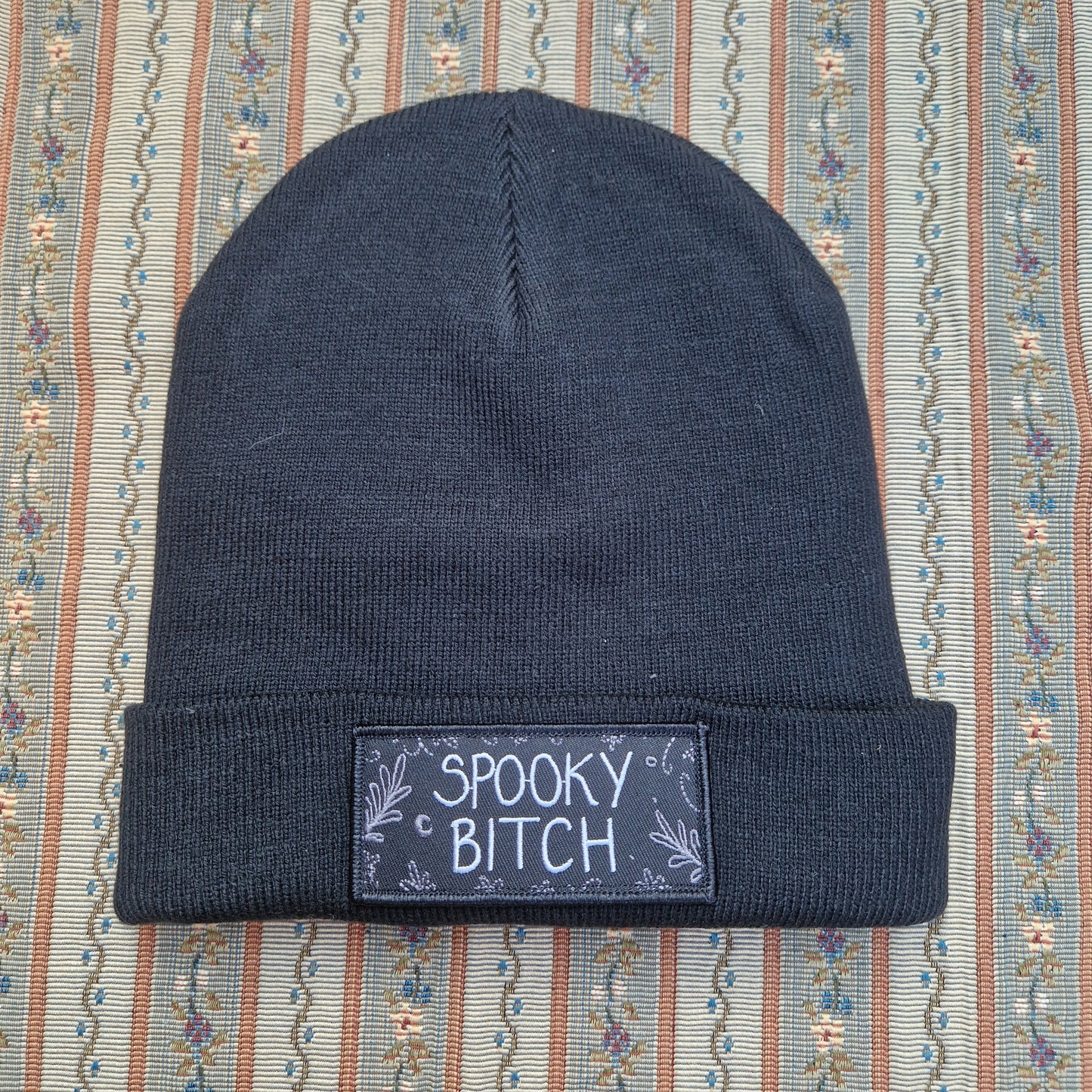 A black beanie sits on a vertical striped textured fabric background. The beanie has a folded up cuff with a rectangular patch on it that has the words 'Spooky Bitch' in white across the middle and grey leafy fronds and crescent moons coming in from the outside edge.