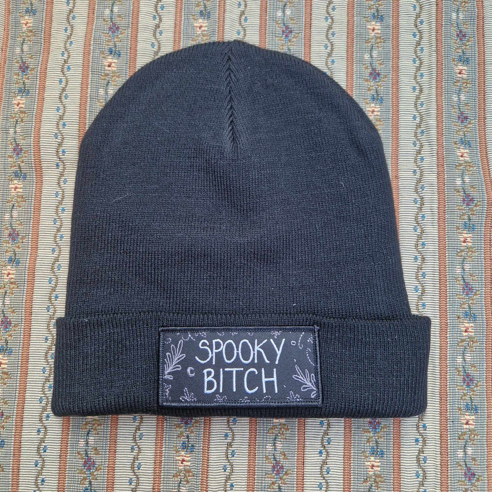 A black beanie sits on a vertical striped textured fabric background. The beanie has a folded up cuff with a rectangular patch on it that has the words 'Spooky Bitch' in white across the middle and grey leafy fronds and crescent moons coming in from the outside edge.