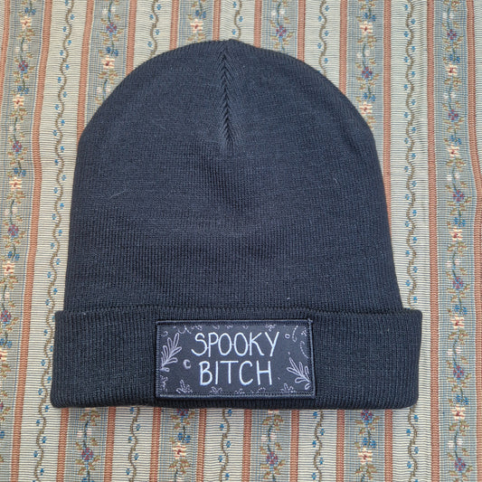 A black beanie sits on a vertical striped textured fabric background. The beanie has a folded up cuff with a rectangular patch on it that has the words 'Spooky Bitch' in white across the middle and grey leafy fronds and crescent moons coming in from the outside edge.
