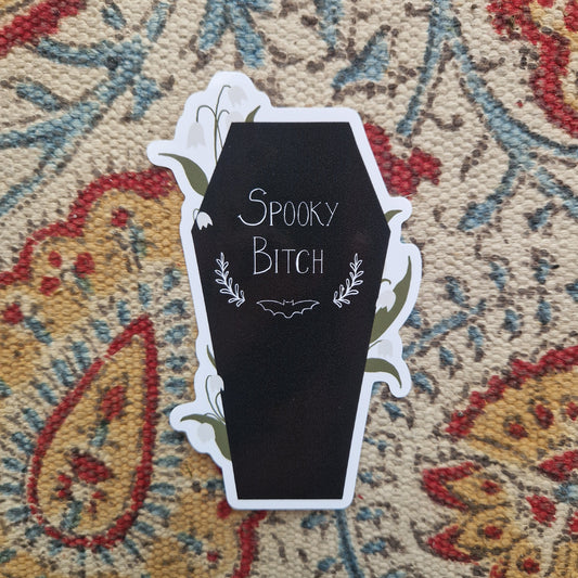 A black coffin shaped sticker with the words 'SPOOKY BITCH' in white with a bat and leafy vines underneath the writing. Around the edge of the coffin are lily of the valley flowering plants. Behind the sticker is a yellow, red, blue and cream patterned fabric. 