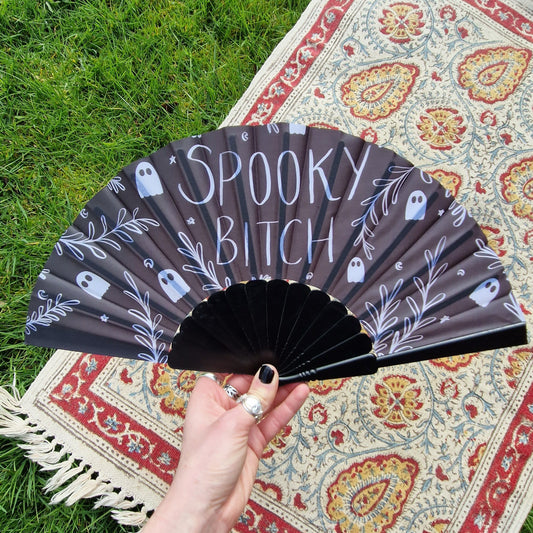 A white hand wearing black nail polish holds a black fan with a white design on it that reads 'SPOOKY BITCH' surrounded by ghosts, leafy vines and starts and crescent moons. Behind the fan is a grassy background and a patterned rug.