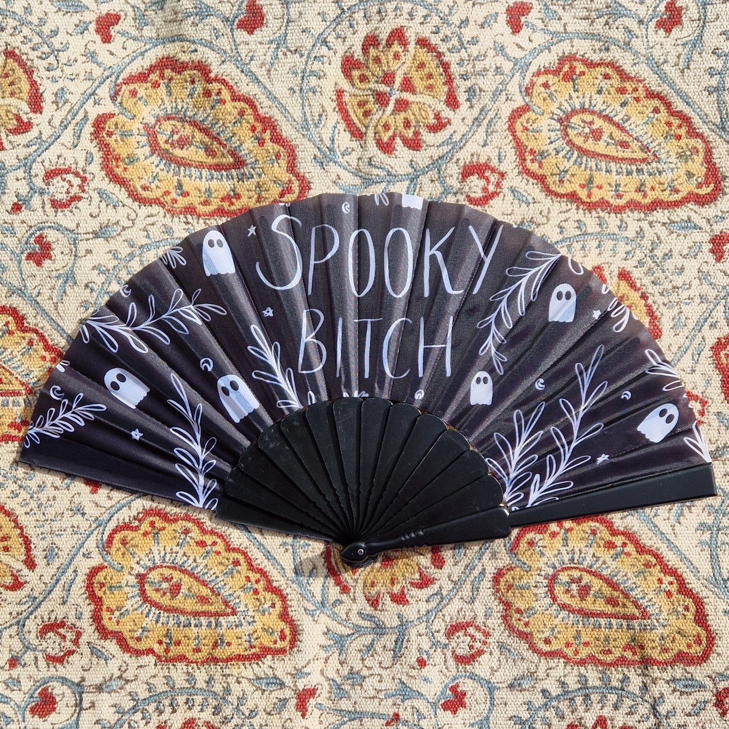 A black fan with the words 'SPOOKY BITCH' written in white handwriting across it and ghosts, leafy vines, stars and crescent moons. Behind the fan is a patterned rug.