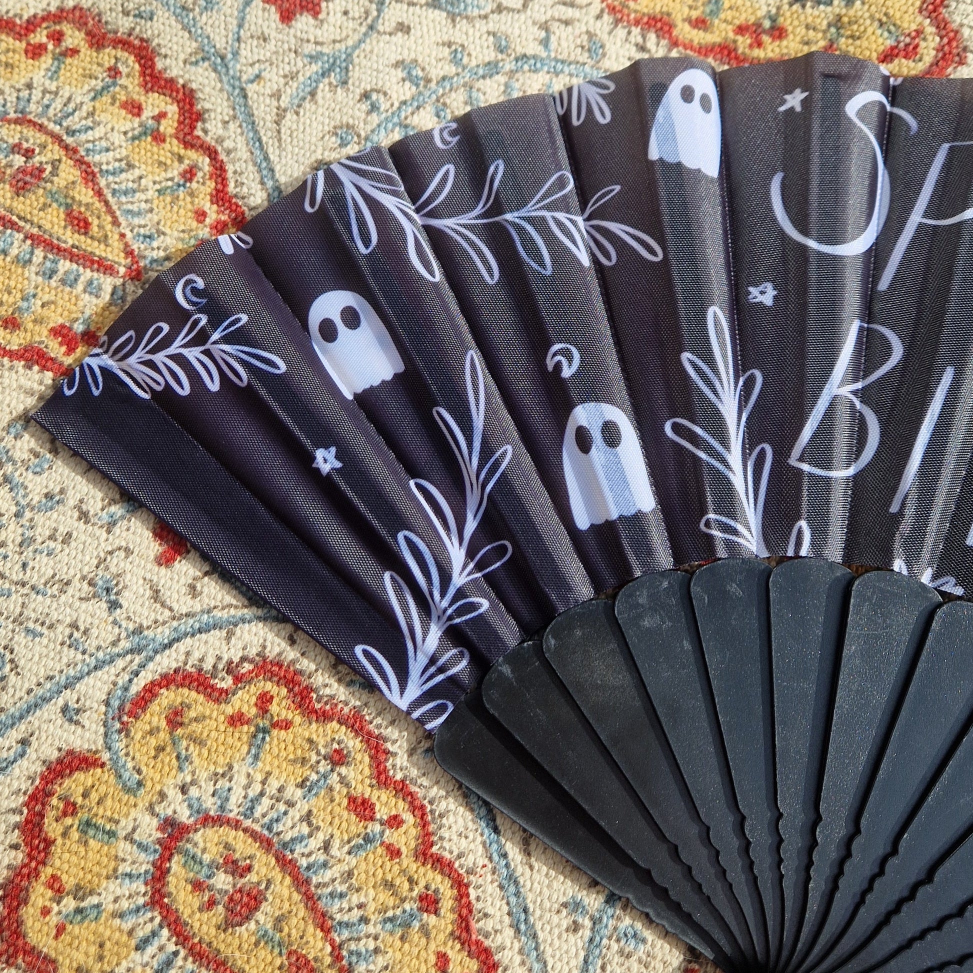 A close up of the left hand side of the black fan showing part of the 'SPOOKY BITCH' writing as well as the leafy vines, ghosts, crescent moons and stars in the design. Behind the fan is a patterned rug.
