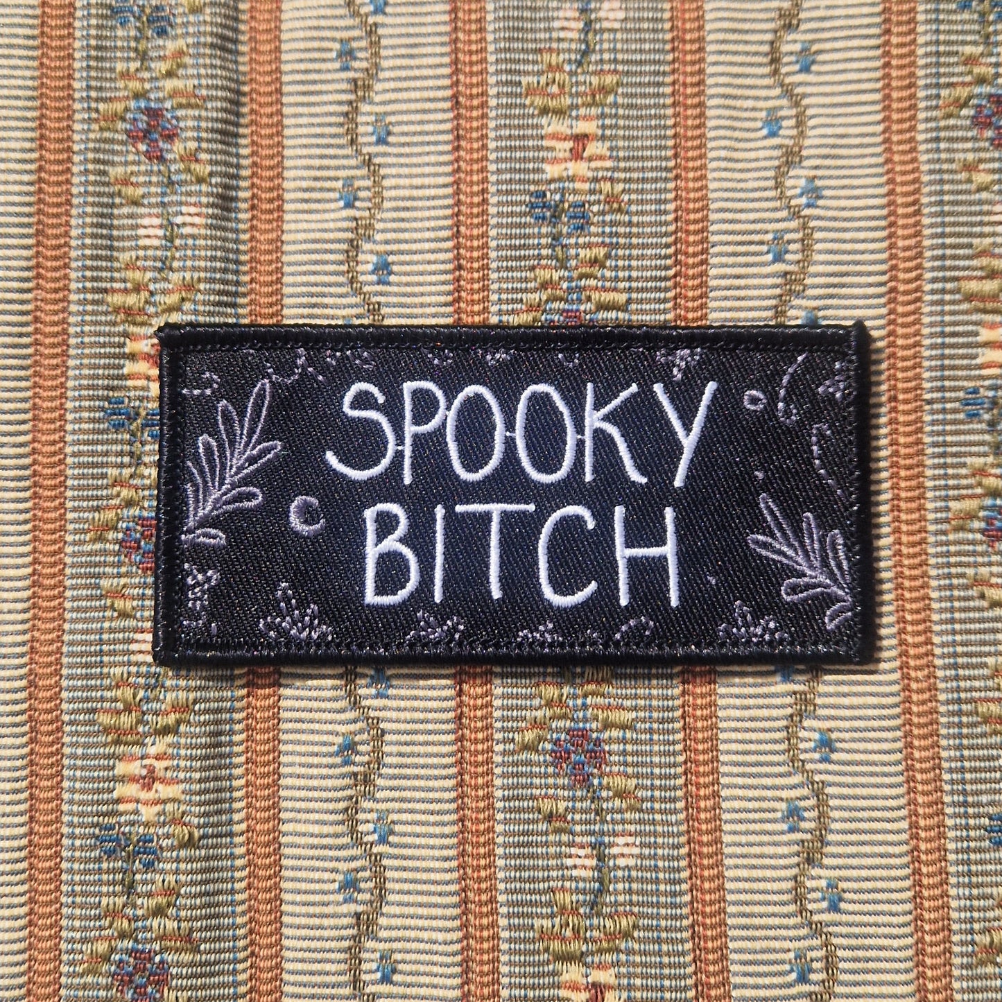 A black rectangular embroidered patch featuring the words 'SPOOKY BITCH' in white writing with grey leafy fronds and a crescent moon around the edges.