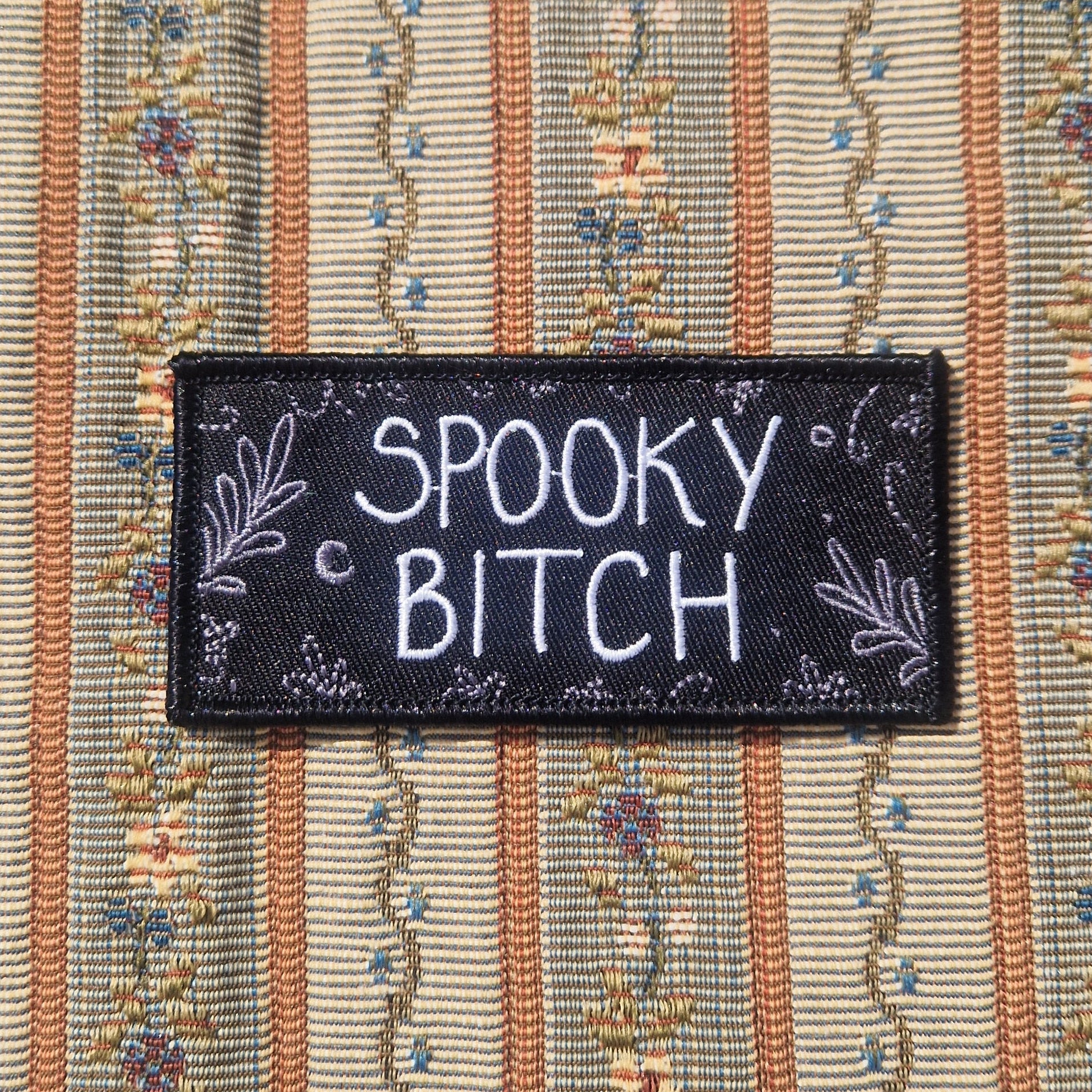 A black rectangular embroidered patch featuring the words 'SPOOKY BITCH' in white writing with grey leafy fronds and a crescent moon around the edges.