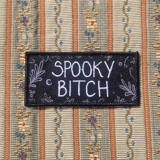 A black rectangular embroidered patch featuring the words 'SPOOKY BITCH' in white writing with grey leafy fronds and a crescent moon around the edges.