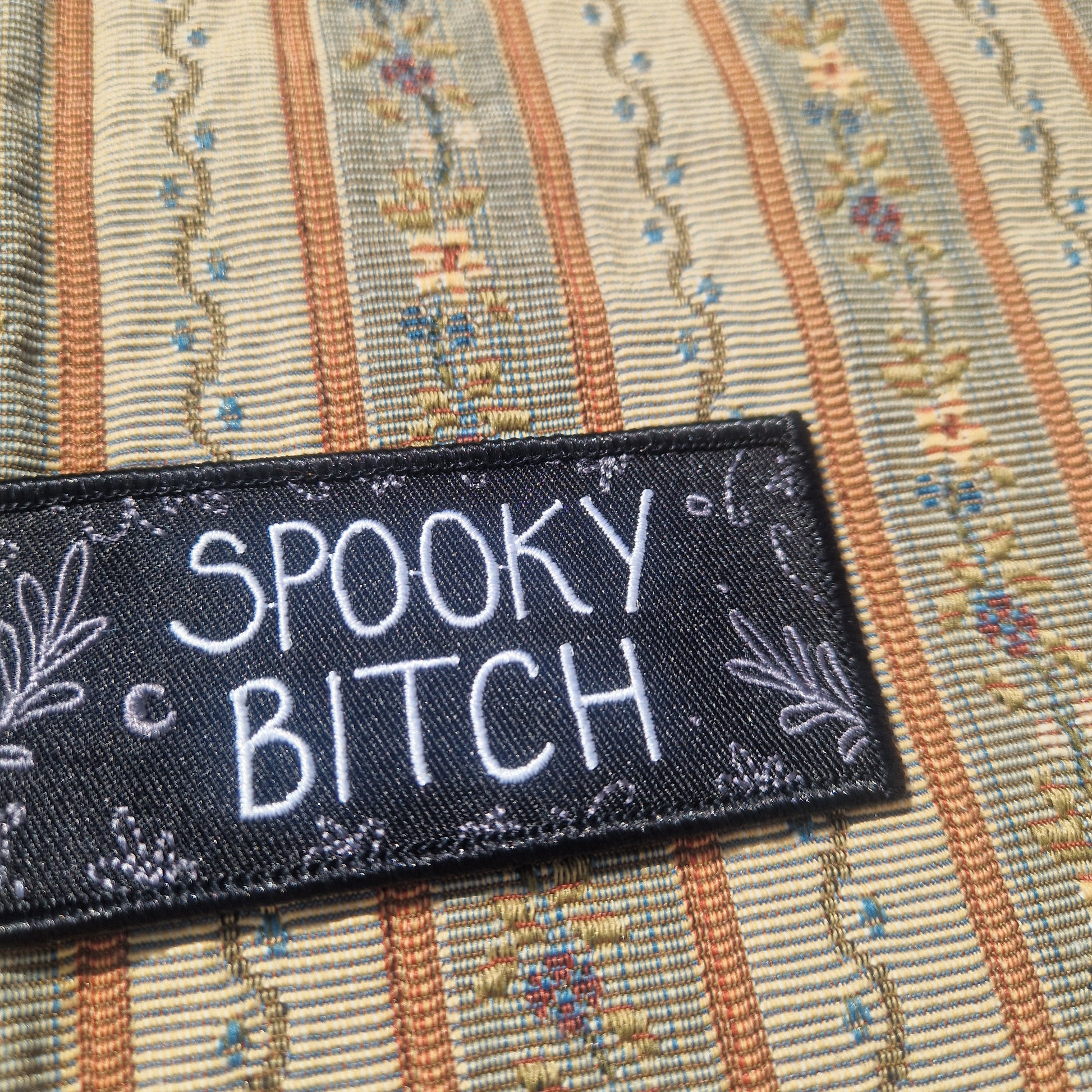 A closer look at the rectangular black 'SPOOKY BITCH' patch, showing off the grey leafy vine and crescent moon design around the edges.